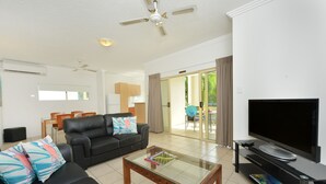 A living room with a black leather sofa, a glass coffee table, a flat-screen TV, and a ceiling fan. - Central Plaza Port Douglas Apartments (Port Douglas)