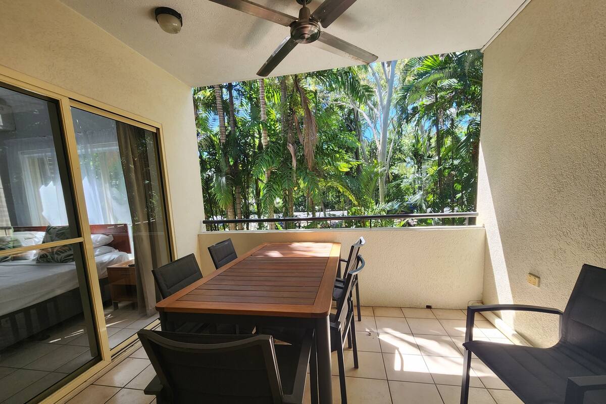 Ein Zimmer mit einem Holzespeisetisch, schwarzen Stühlen, einem Deckenventilator und einem großen Fenster mit Blick auf tropische Bäume.