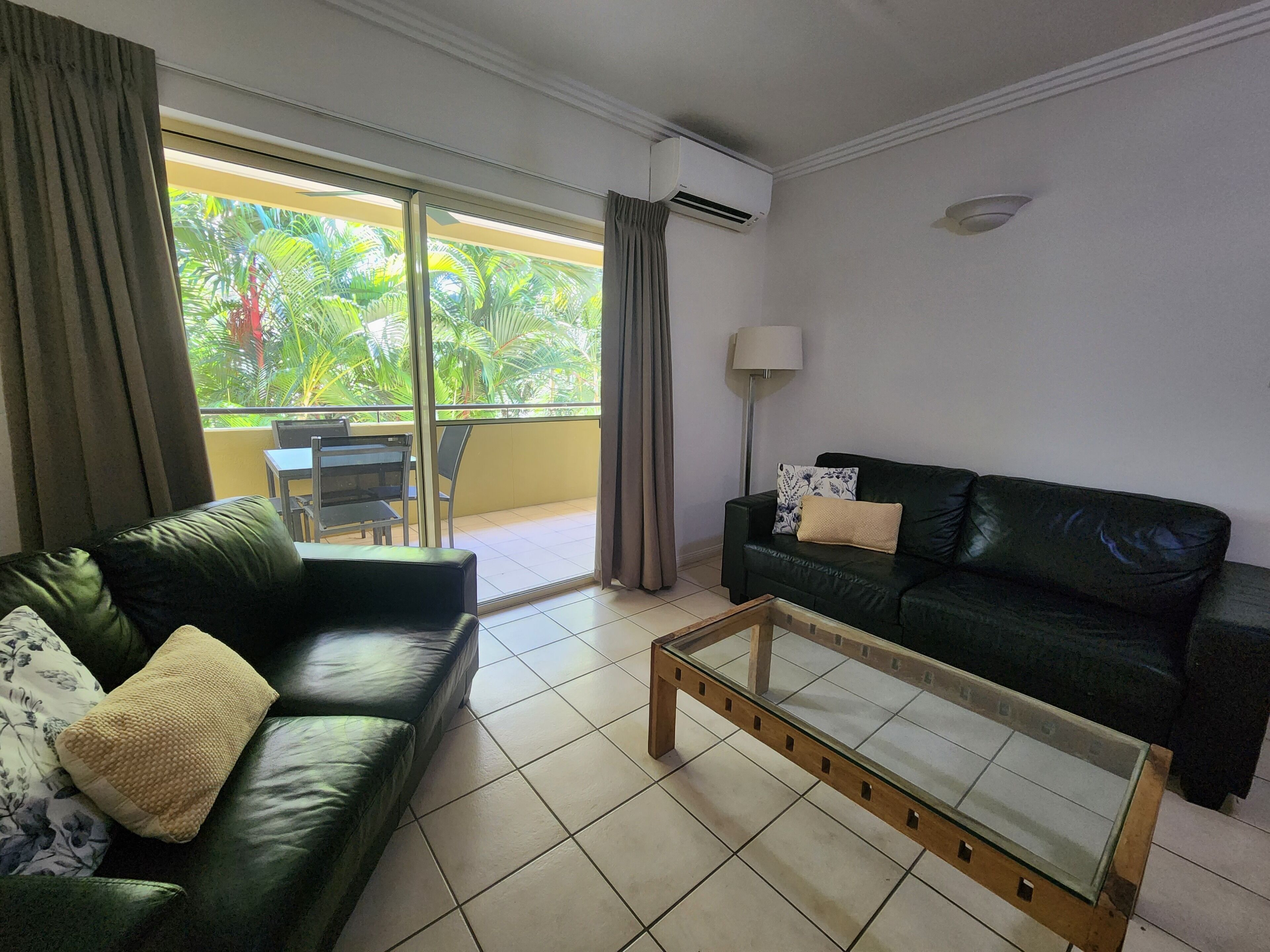 Een woonkamer met een groene leren bank, een glazen salontafel, een zwarte leren bank, een eettafel met stoelen en uitzicht op groen via een schuifdeur.