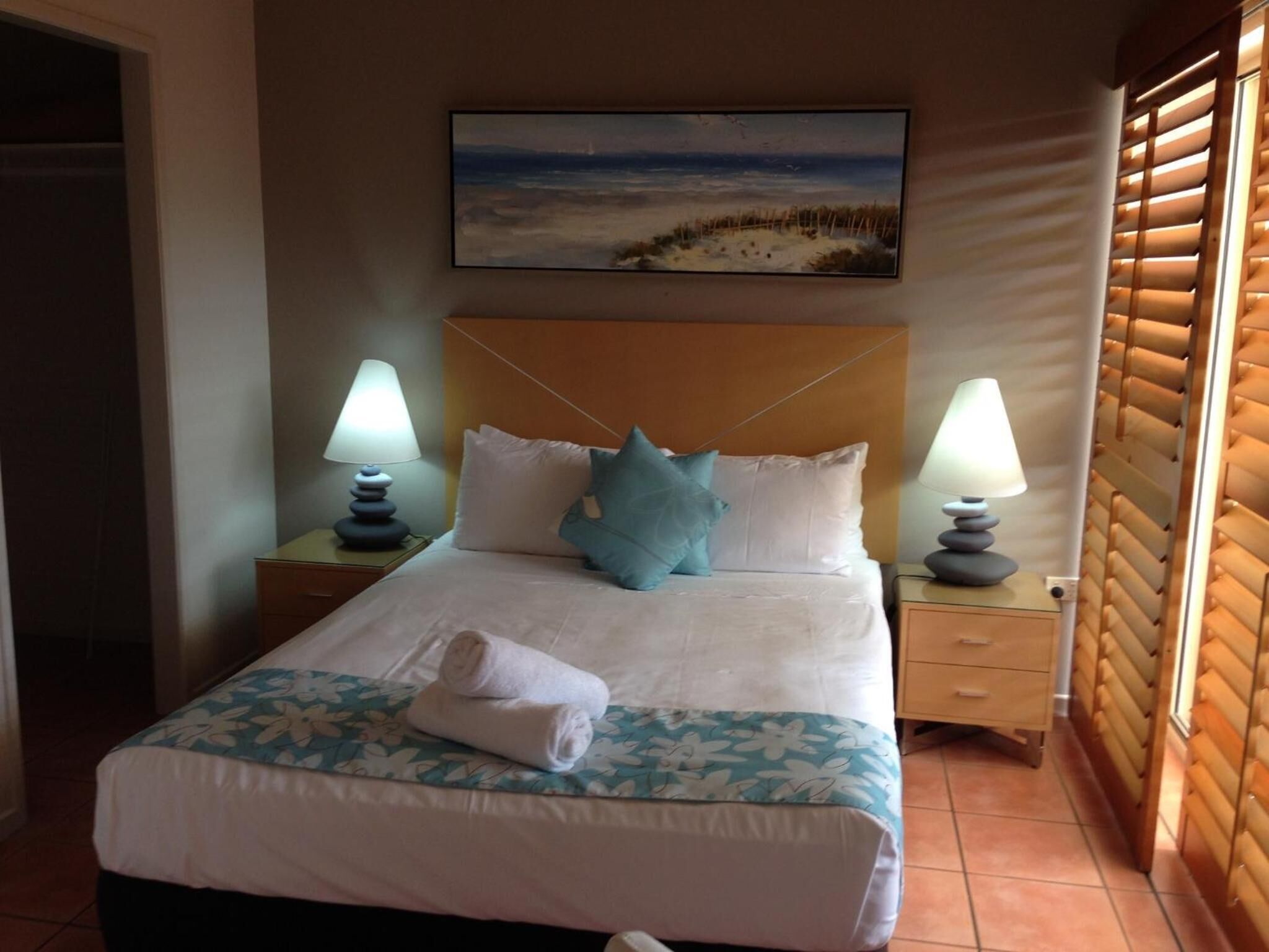 A hotel room with a bed, two bedside lamps, a painting of a beach scene, and a window with blinds.