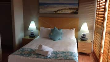 Habitación de hotel con cama, dos lámparas de noche, un cuadro de una playa y una ventana con persianas.