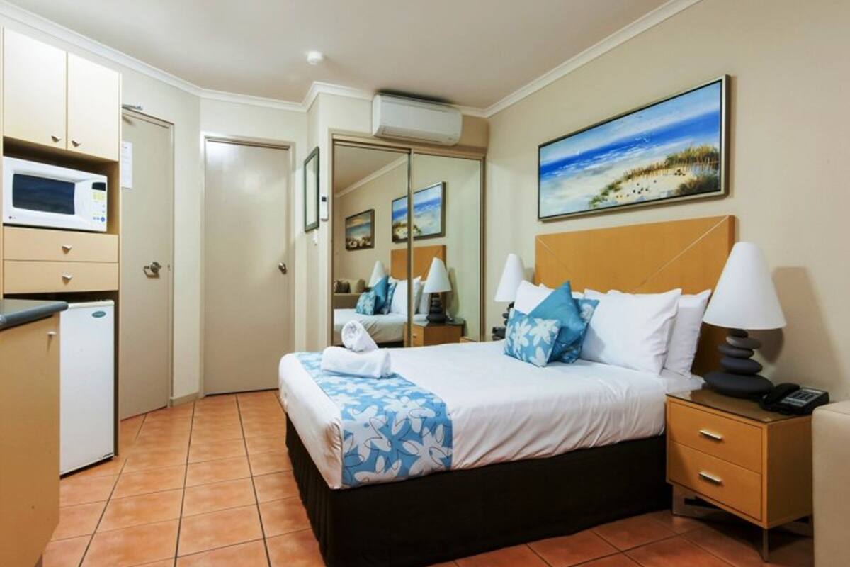 A hotel room with a bed, bedside tables, a microwave, a refrigerator, a painting of a beach, and a tiled floor.