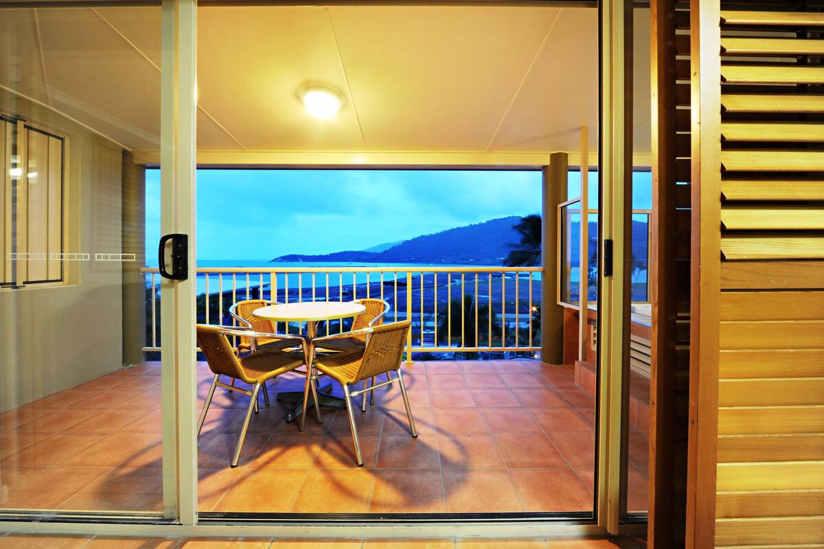 A balcony with a table and chairs, a view of the mountains, and a glass door leading to the interior.