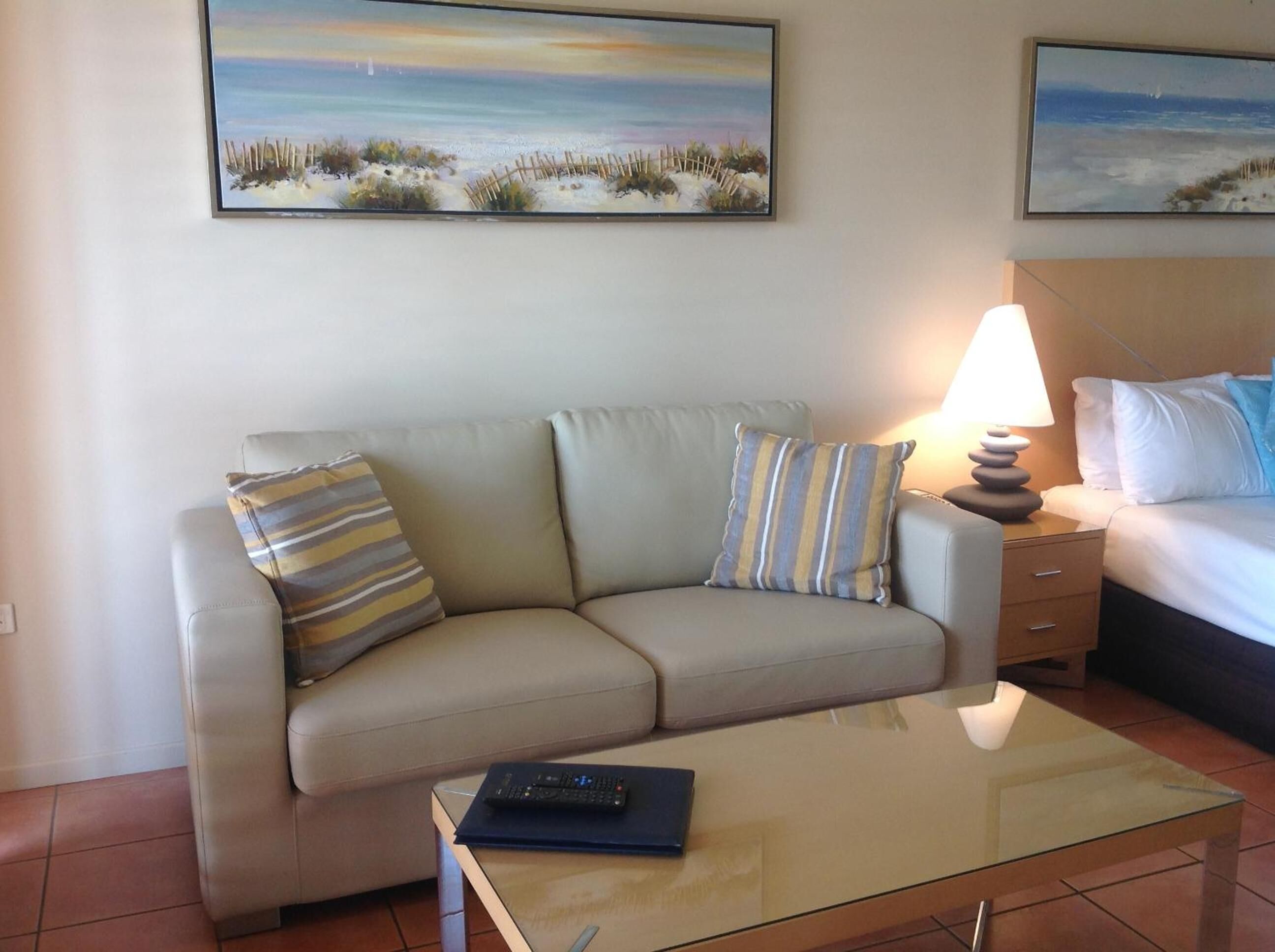 A hotel room with a beige sofa, striped pillows, a glass coffee table, a remote control, and a lamp.