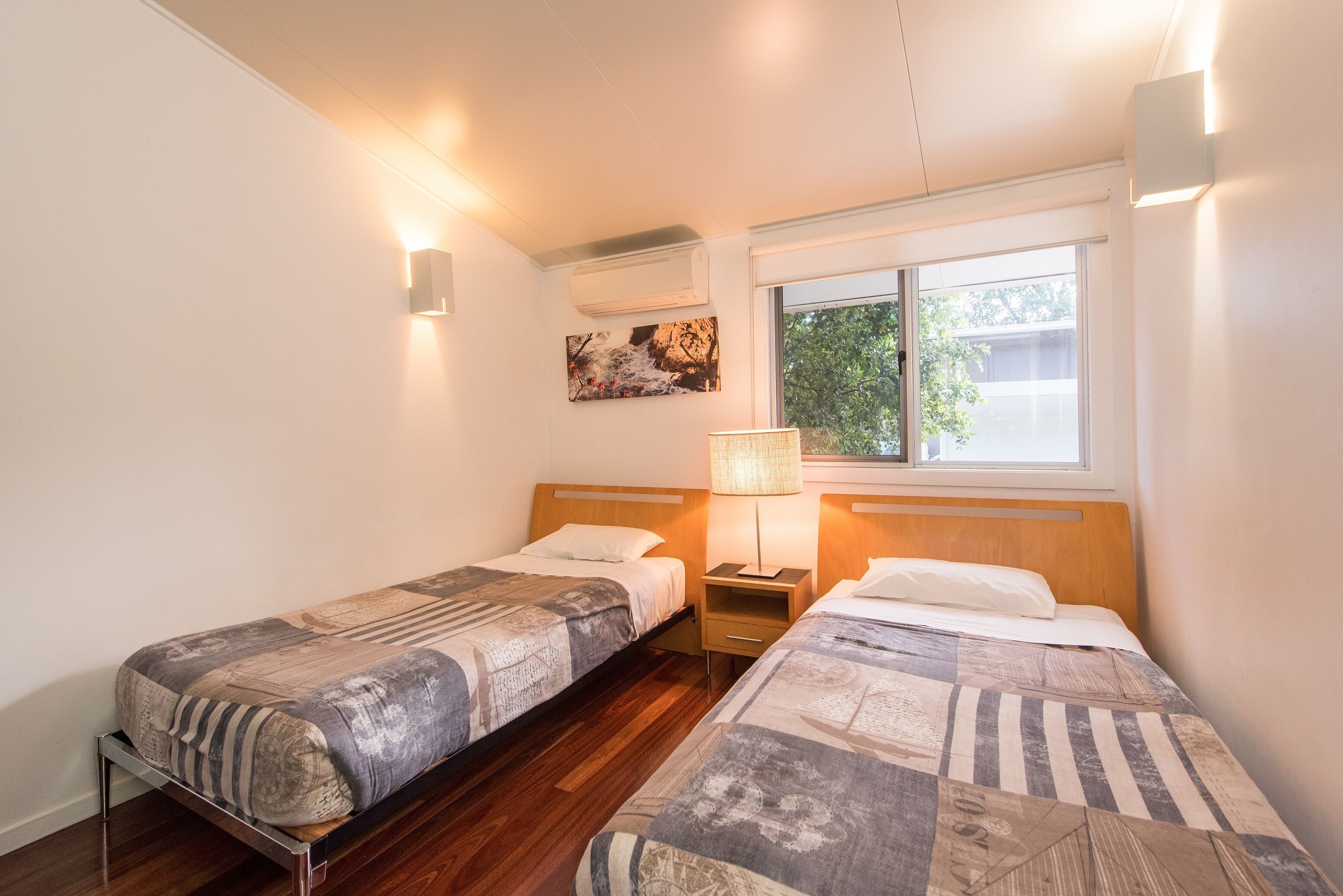 A bedroom with two single beds, a nightstand with a lamp, a window with a view of trees, and an air conditioning unit on the wall.