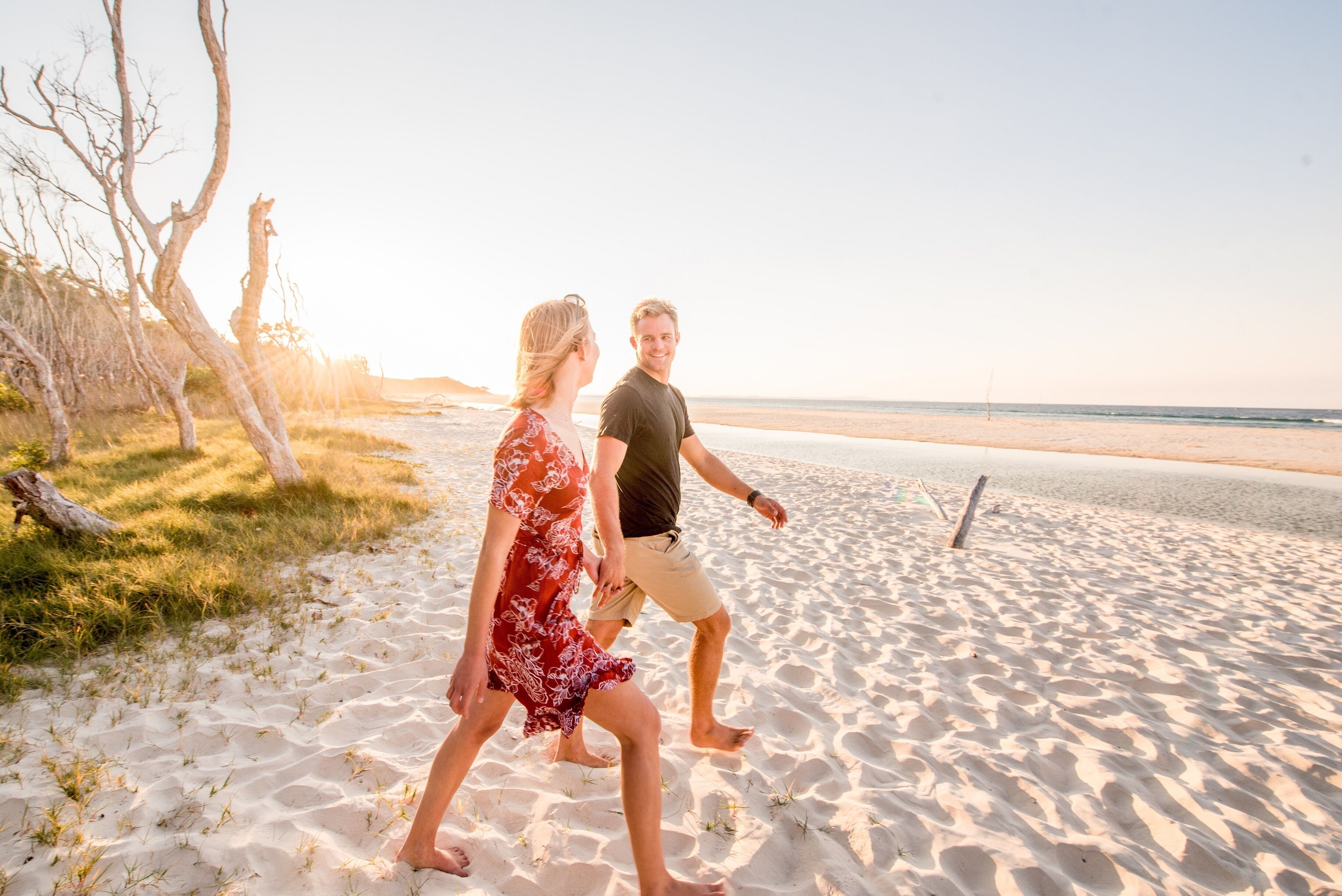 Una pareja caminando sobre una playa arenosa, con cielo despejado y algunos árboles.