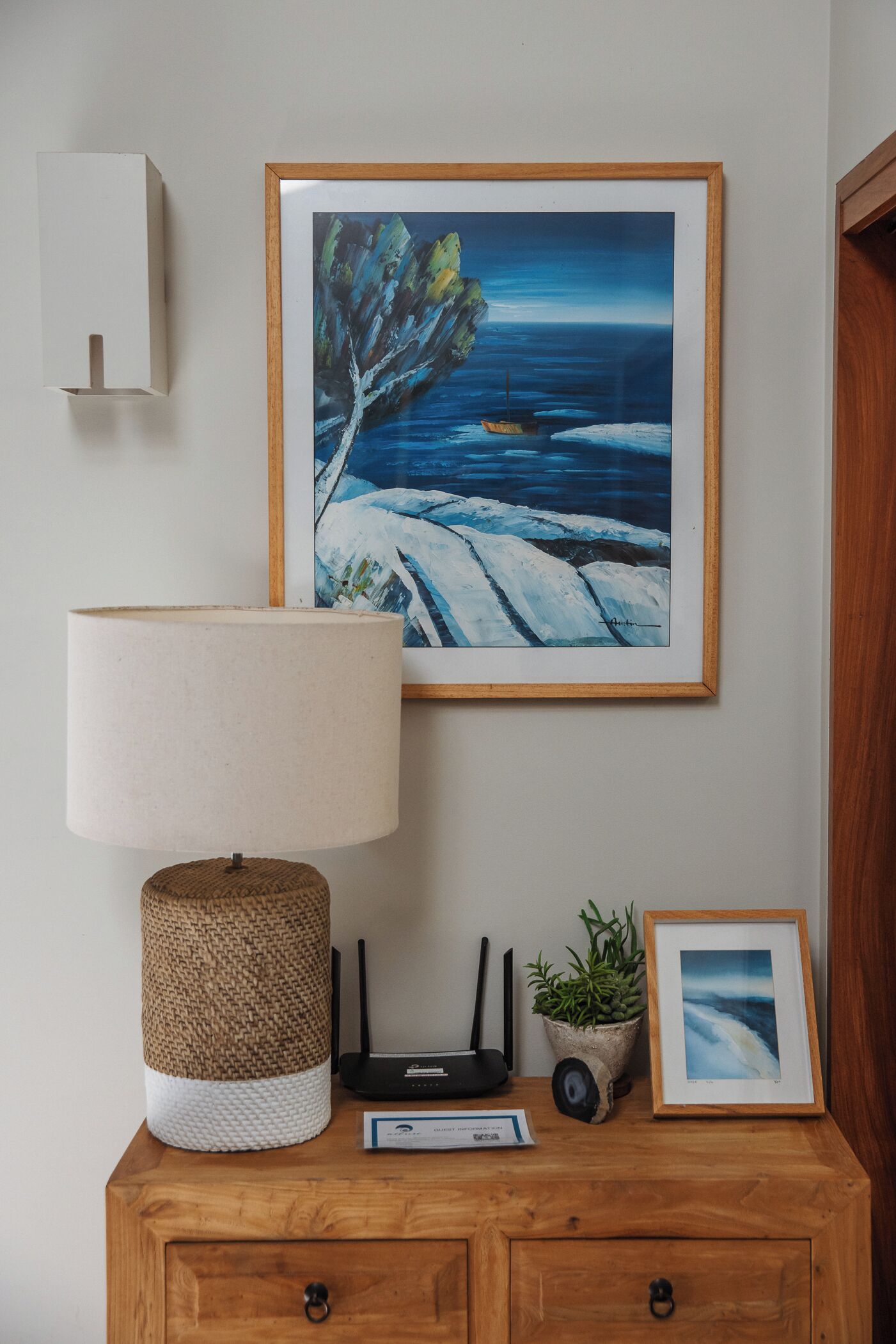 A wooden nightstand with a lamp, a framed painting of a coastal scene, a small potted plant, and a framed photograph.