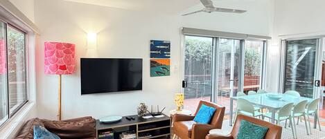 A modern living room with a flat-screen TV, leather sofas, a dining area with a glass table, and a ceiling fan.