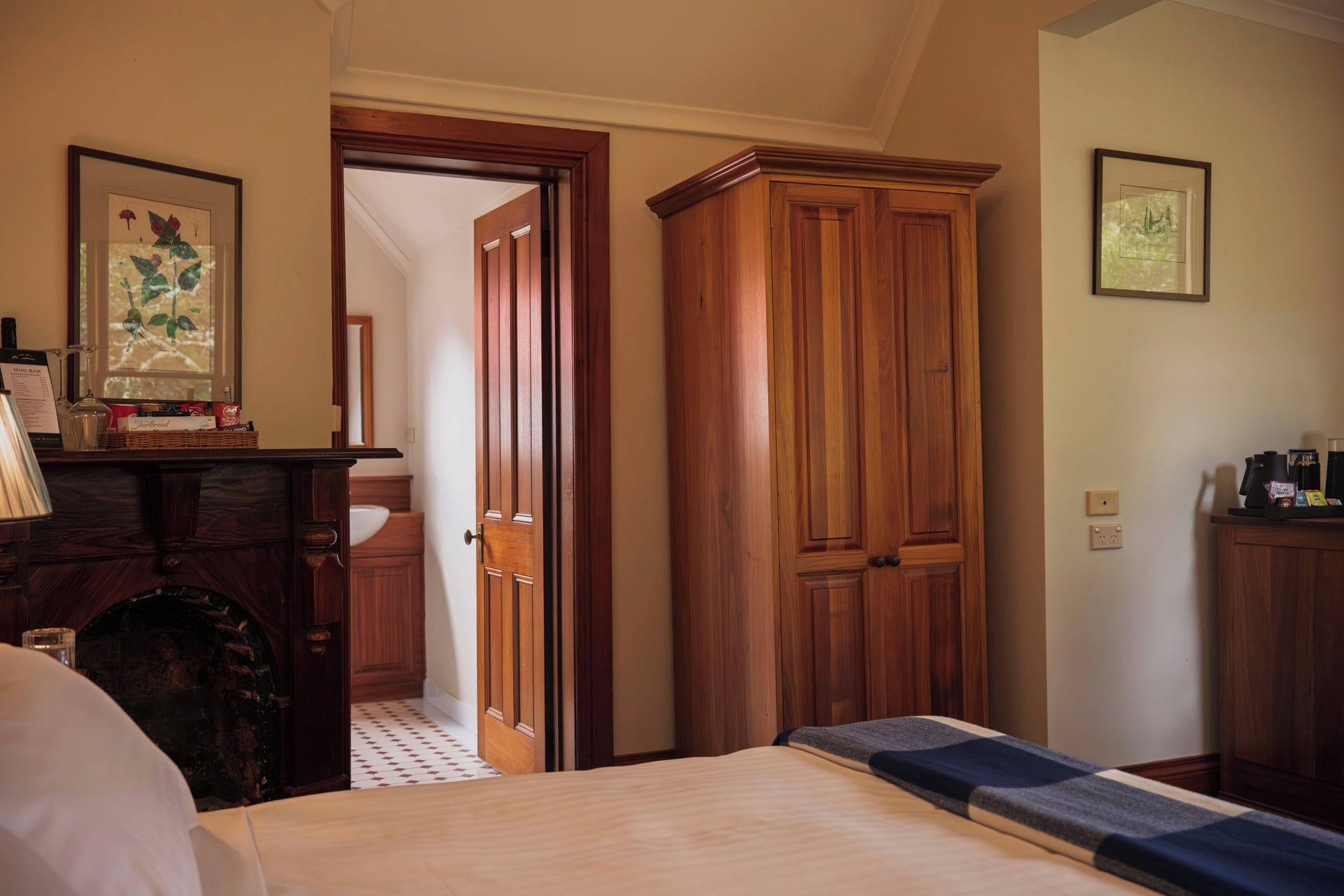 A hotel room with a bed, wooden wardrobe, a desk with a lamp, and a framed botanical print on the wall.