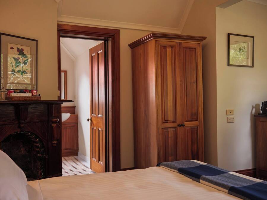 A hotel room with a bed, wooden wardrobe, a desk with a lamp, and a framed botanical print on the wall.