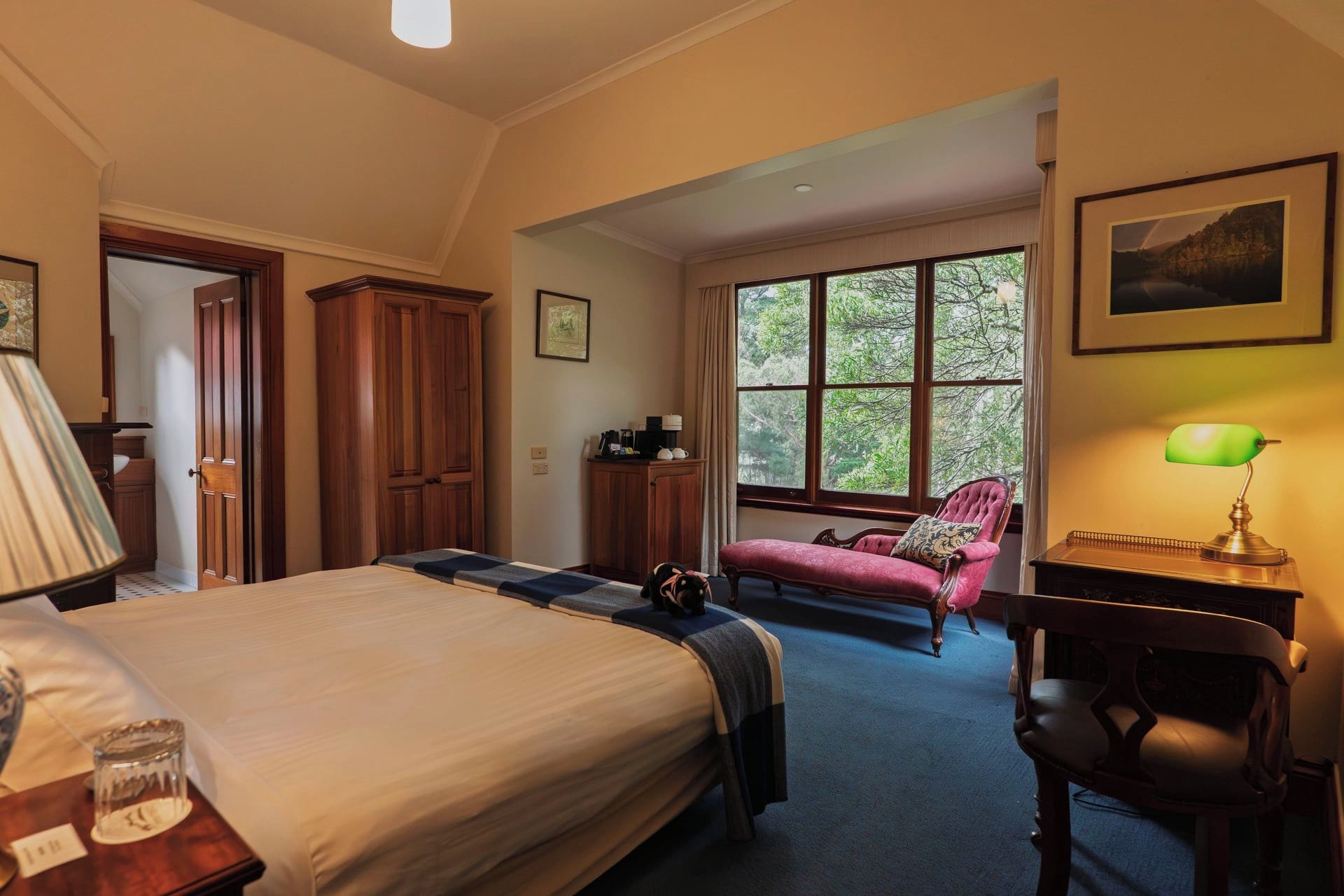 A bedroom with a large bed, a desk with a lamp, a chair, and a window with a view of trees.