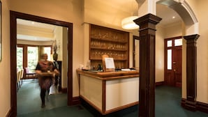 A hotel lobby with a reception desk, a wooden bookshelf with glassware, and a large mirror. - Franklin Manor (Strahan)
