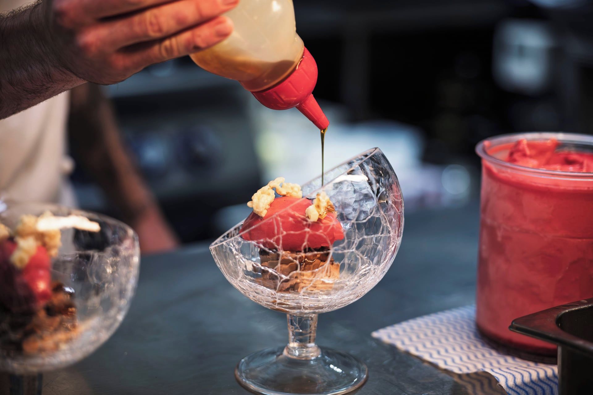 A hand pouring a sauce into a dessert with a red topping, served in a clear glass bowl.