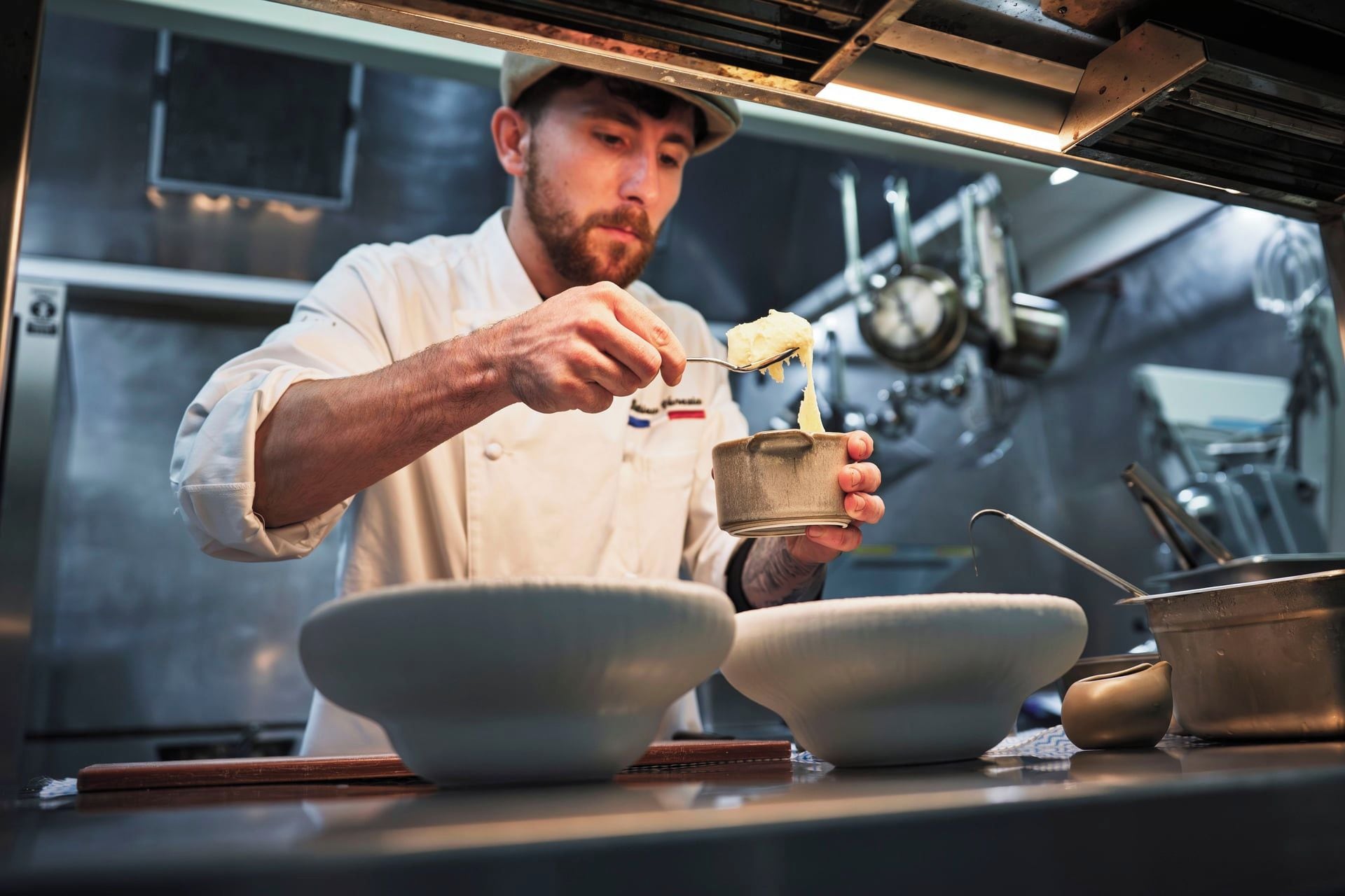 Um chef em uma cozinha profissional, segurando uma tigela e uma colher, com diversos utensílios e equipamentos de cozinha ao fundo.