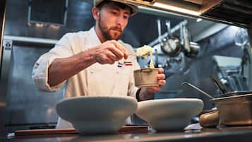 Um chef em uma cozinha profissional, segurando uma tigela e uma colher, com diversos utensílios e equipamentos de cozinha ao fundo.