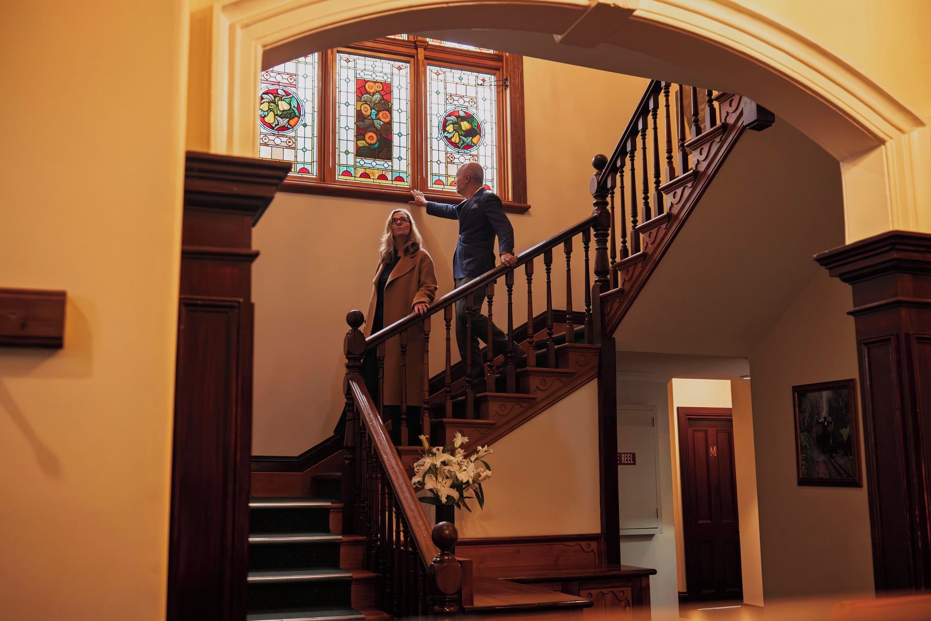 A grand staircase with a wooden handrail and balusters, leading to a landing with a stained glass window above.