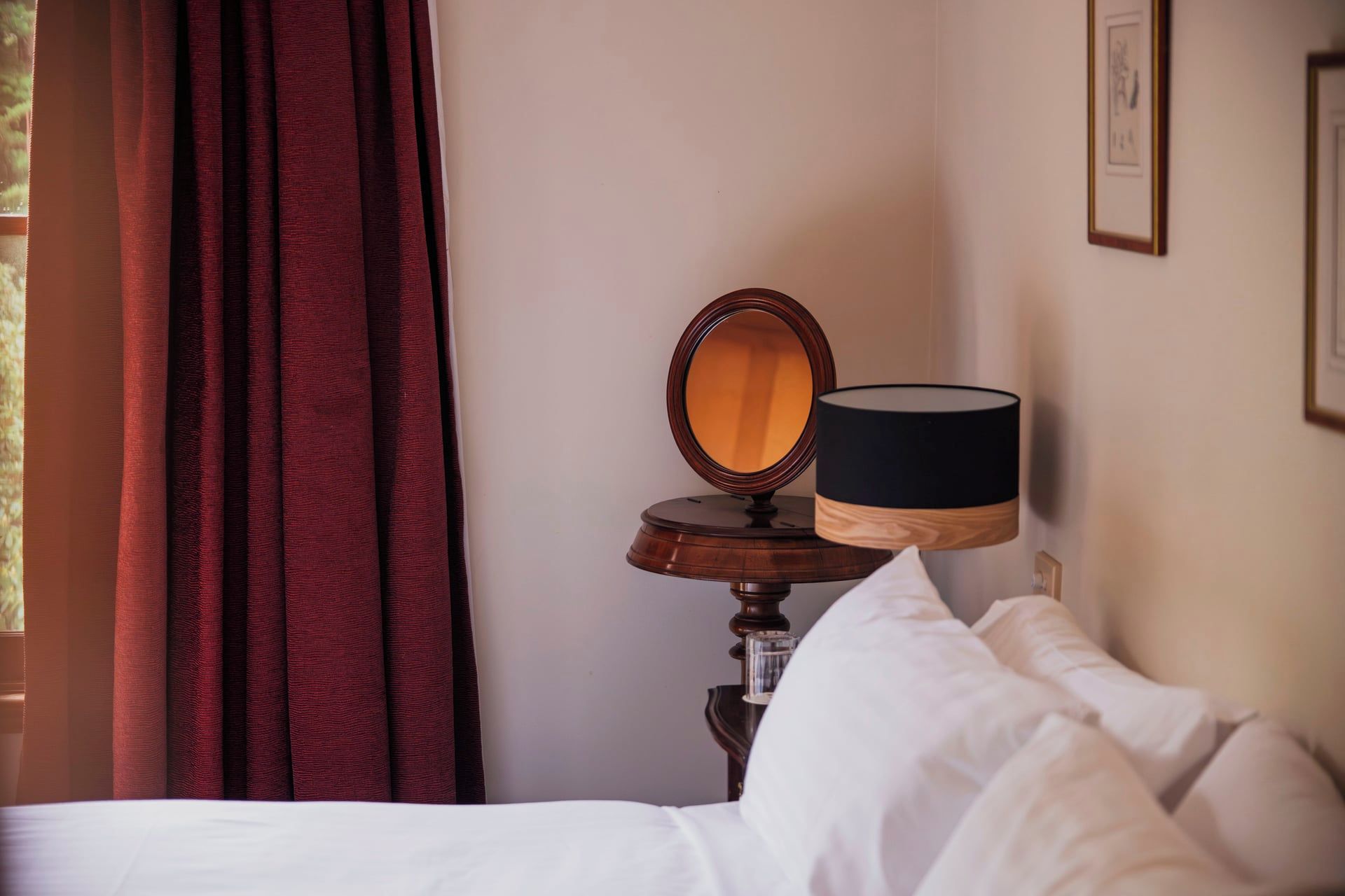 A hotel room with a bed, a bedside table with a lamp, a mirror, and a window with curtains.