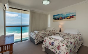 A hotel room with two beds, a desk, a chair, a painting of boats on the wall, and a view of the ocean. - Norfolk Luxury Beachfront Apartments (Main Beach)