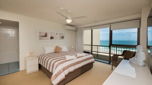 Une chambre d’hôtel avec un lit, une table de chevet, un ventilateur de plafond, une grande fenêtre donnant sur la plage et une statue posée sur le rebord de la fenêtre.