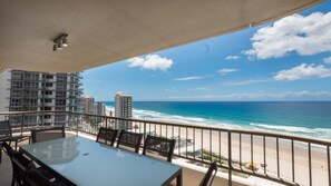 A balcony with a glass table and chairs, overlooking a beach and ocean with buildings in the background. - Norfolk Luxury Beachfront Apartments (Main Beach)