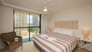 A hotel room with a large bed, a ceiling fan, a view of high-rise buildings, and a wicker chair.