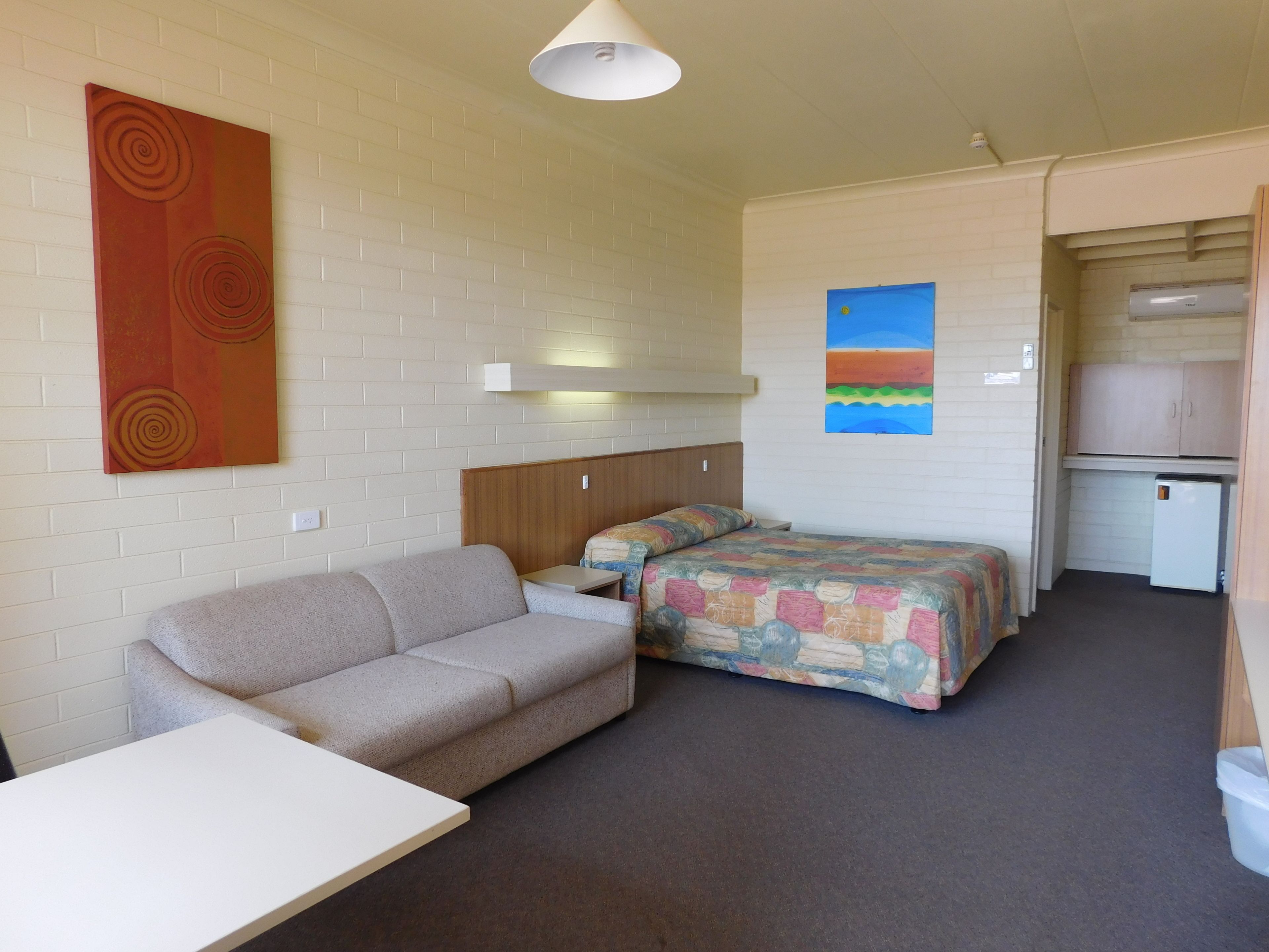 A compact room with a sofa, a bed with a colorful quilt, a small table, a wall art piece, and a kitchenette area.