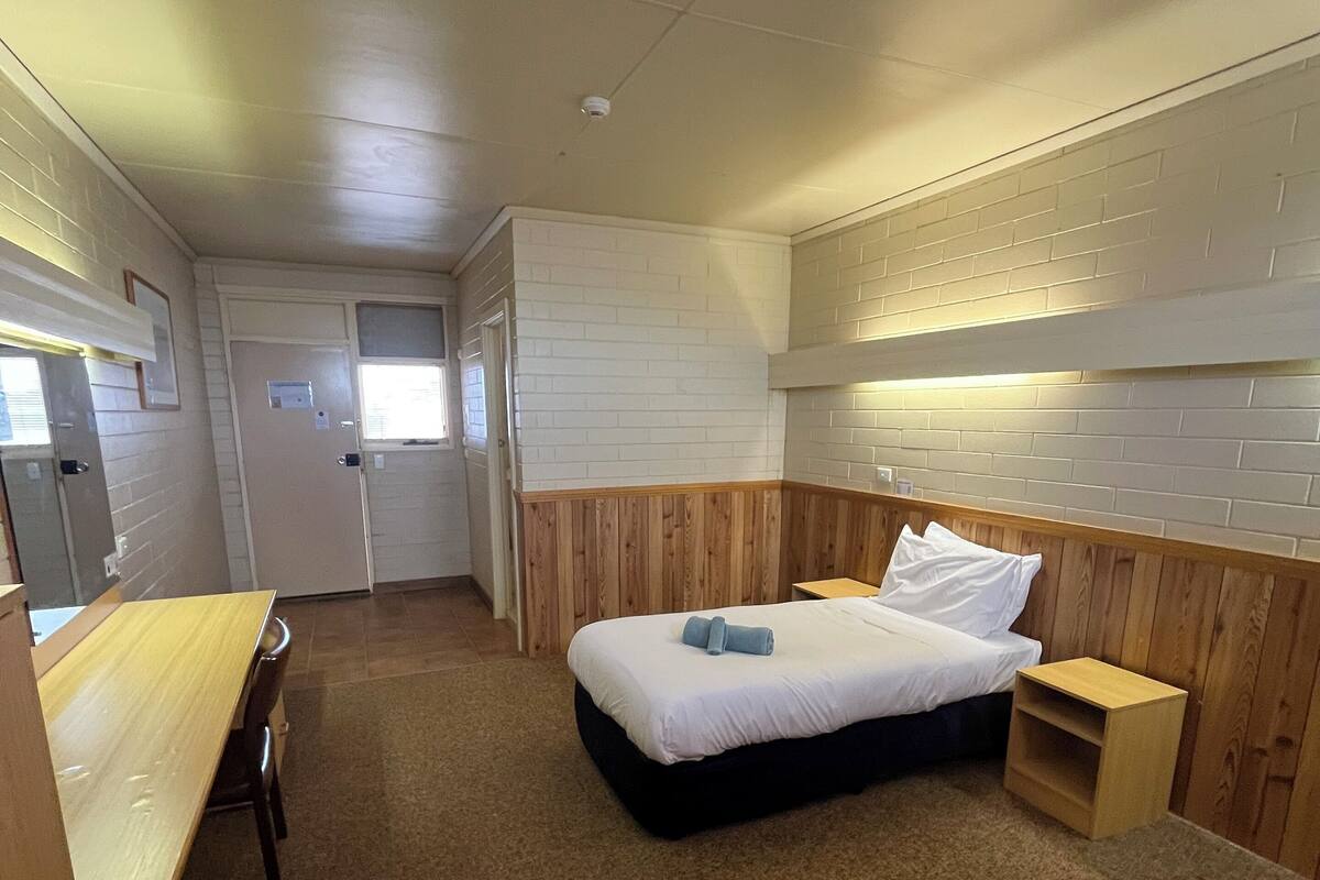 A compact hotel room with a single bed, a small bedside table, a wooden bench, and a desk with a chair.