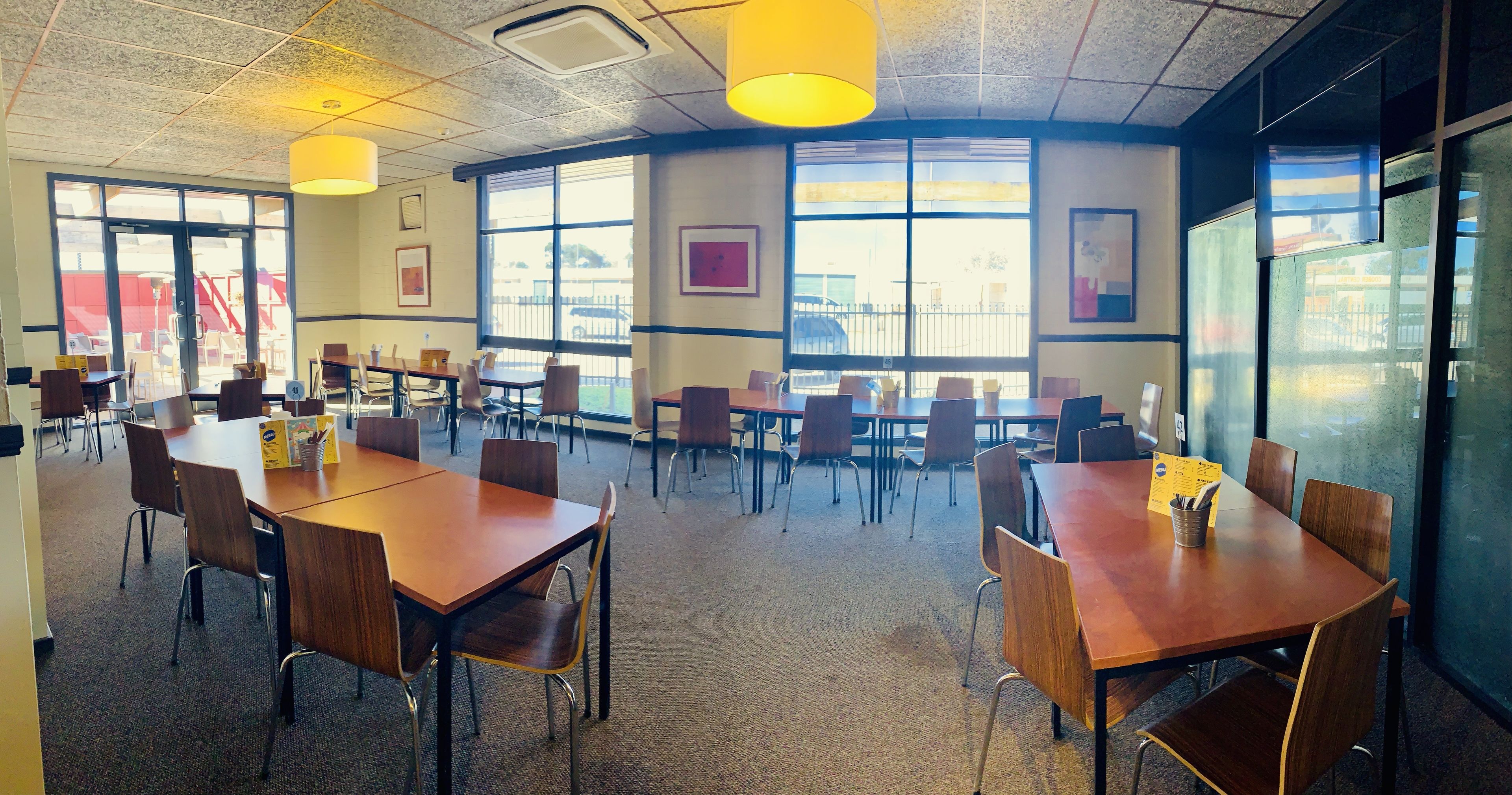 Un comedor interior amplio con múltiples mesas y sillas de madera, ventanas grandes que proporcionan luz natural y obras de arte enmarcadas en las paredes.