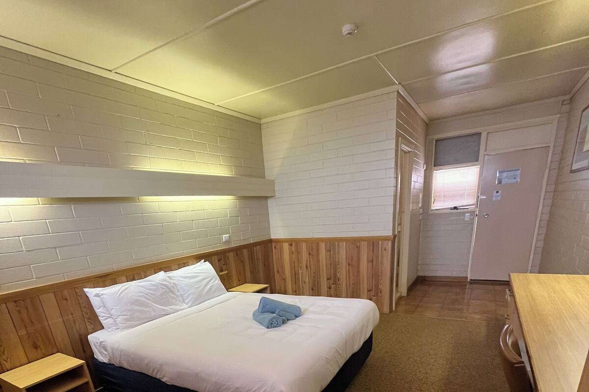A hotel room with a bed, a wooden nightstand, a desk, and a door.