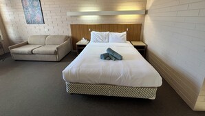Une chambre d’hôtel avec un grand lit, une tête de lit en bois, une table de chevet, un fauteuil beige et une œuvre d’art accrochée au mur.