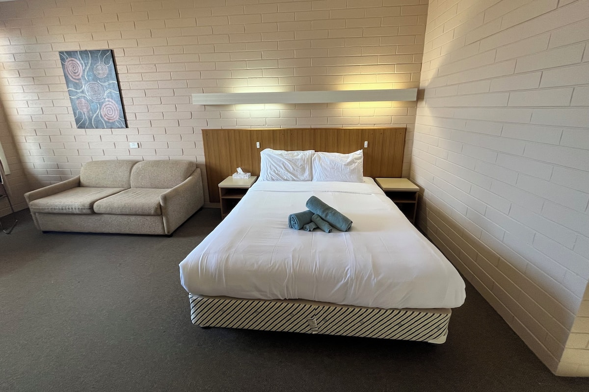A hotel room with a large bed, a wooden headboard, a nightstand, a beige armchair, and a wall-mounted artwork.