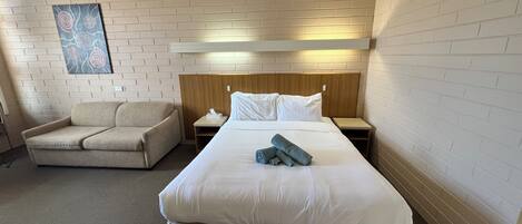 Una habitación de hotel con una cama grande, cabecera de madera, mesita de noche, un sillón beige y una obra de arte colgada en la pared.
