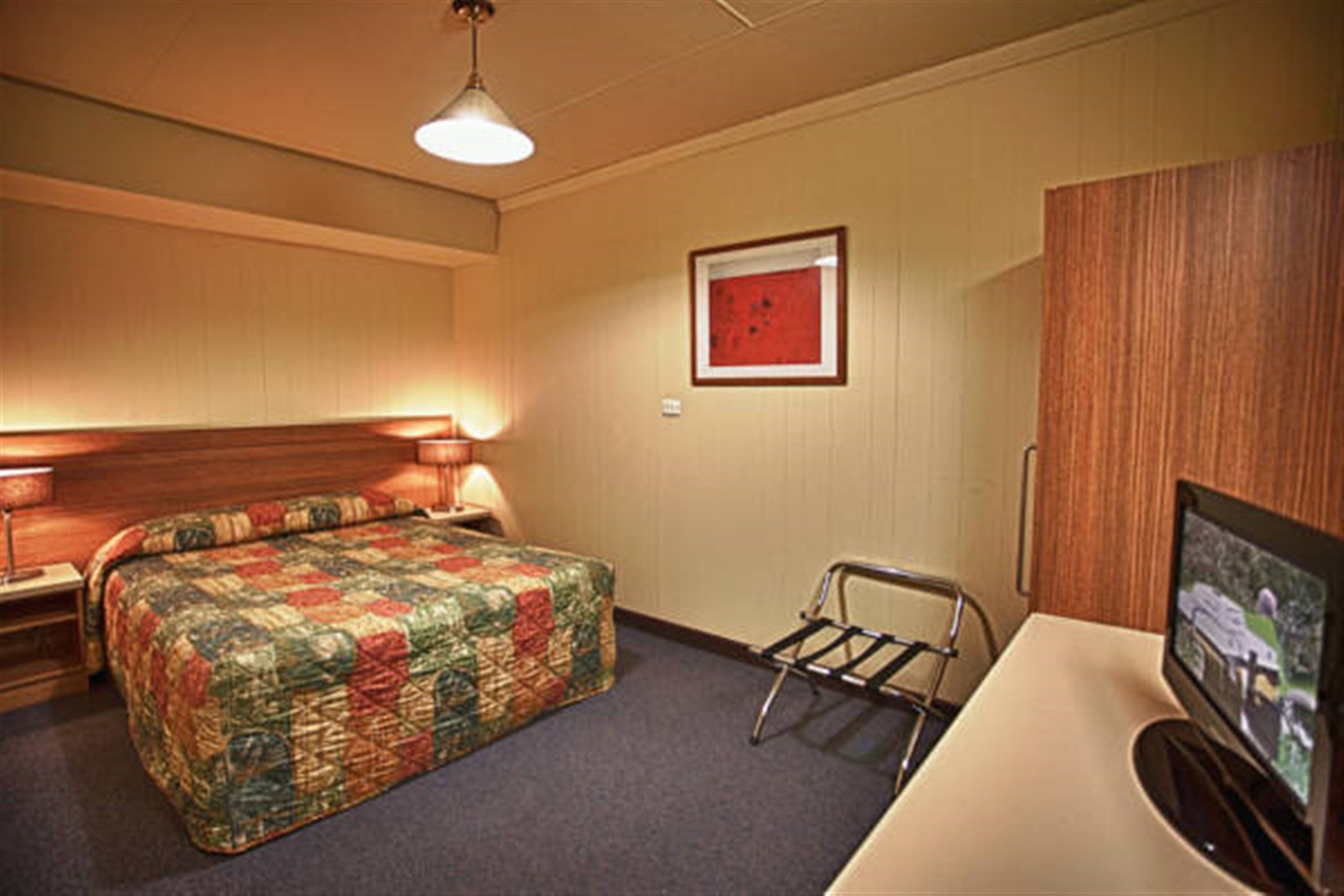 A hotel room with a bed, bedside table, lamp, TV, and a framed picture on the wall.