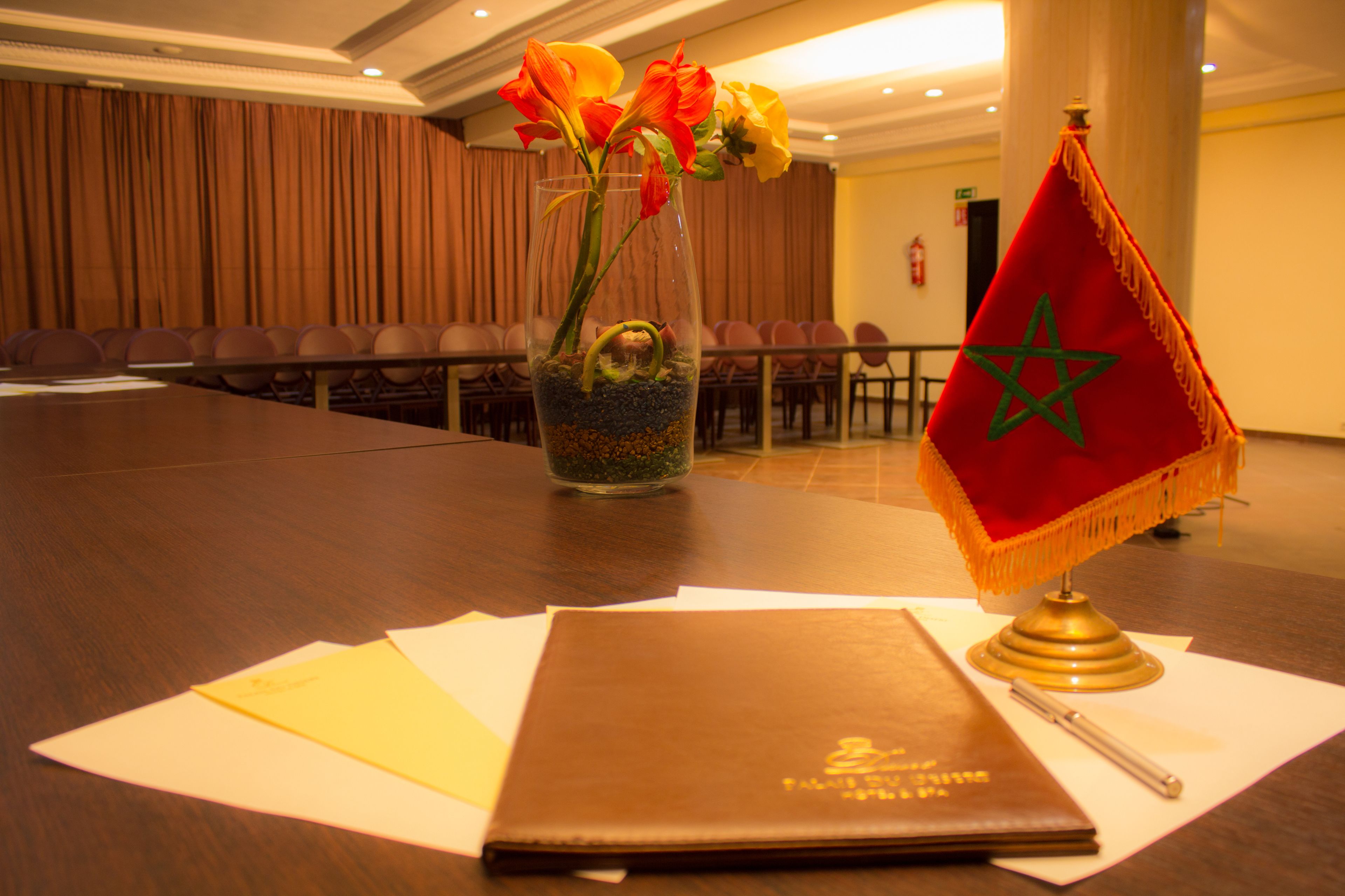 Une salle de conférence avec une table préparée pour une réunion, un vase rempli de fleurs, un drapeau marocain et un ordinateur portable.