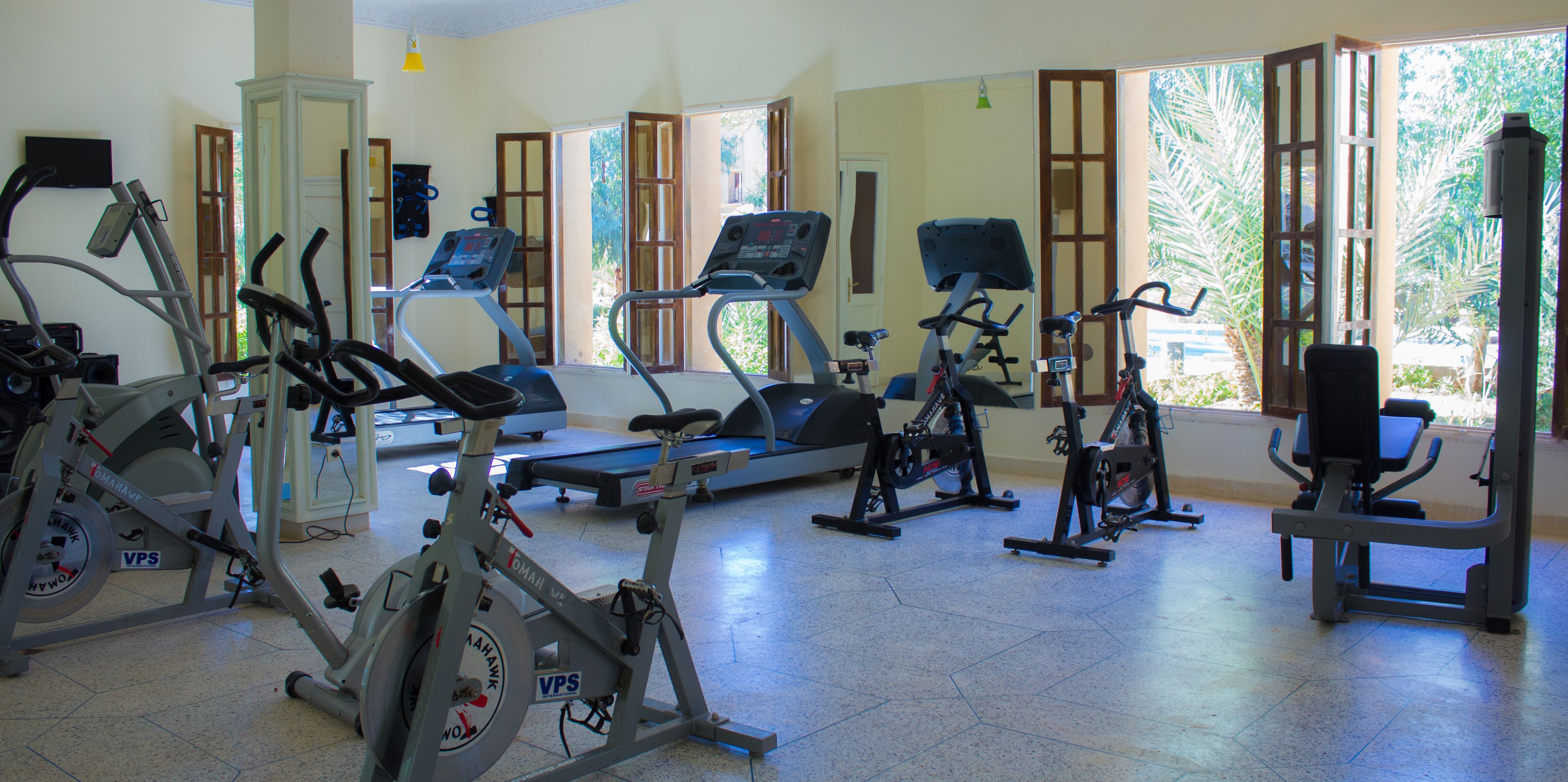 Un gimnasio con diversas máquinas de ejercicio, amplias ventanas y una puerta que da al exterior.