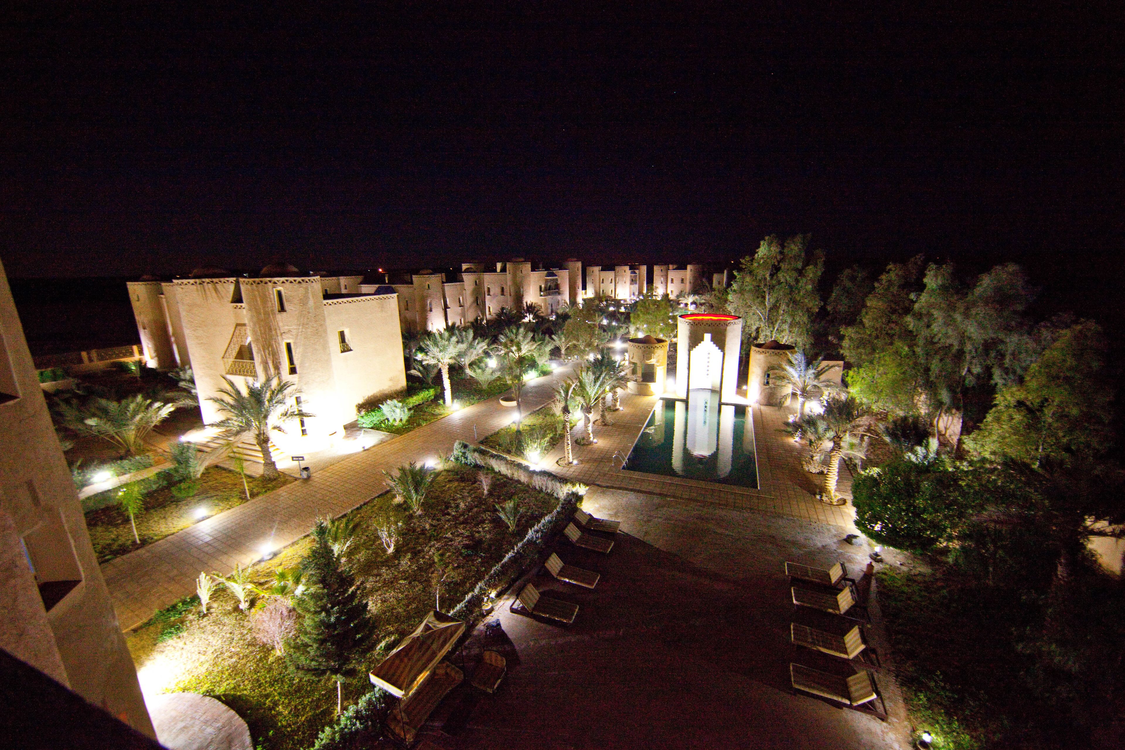Une vue nocturne d’un complexe hôtelier avec une piscine, des palmiers et une allée bien éclairée.