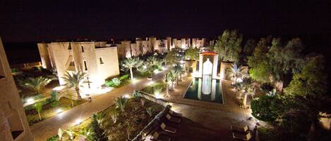 A nighttime view of a resort with a swimming pool, palm trees, and a well-lit walkway.