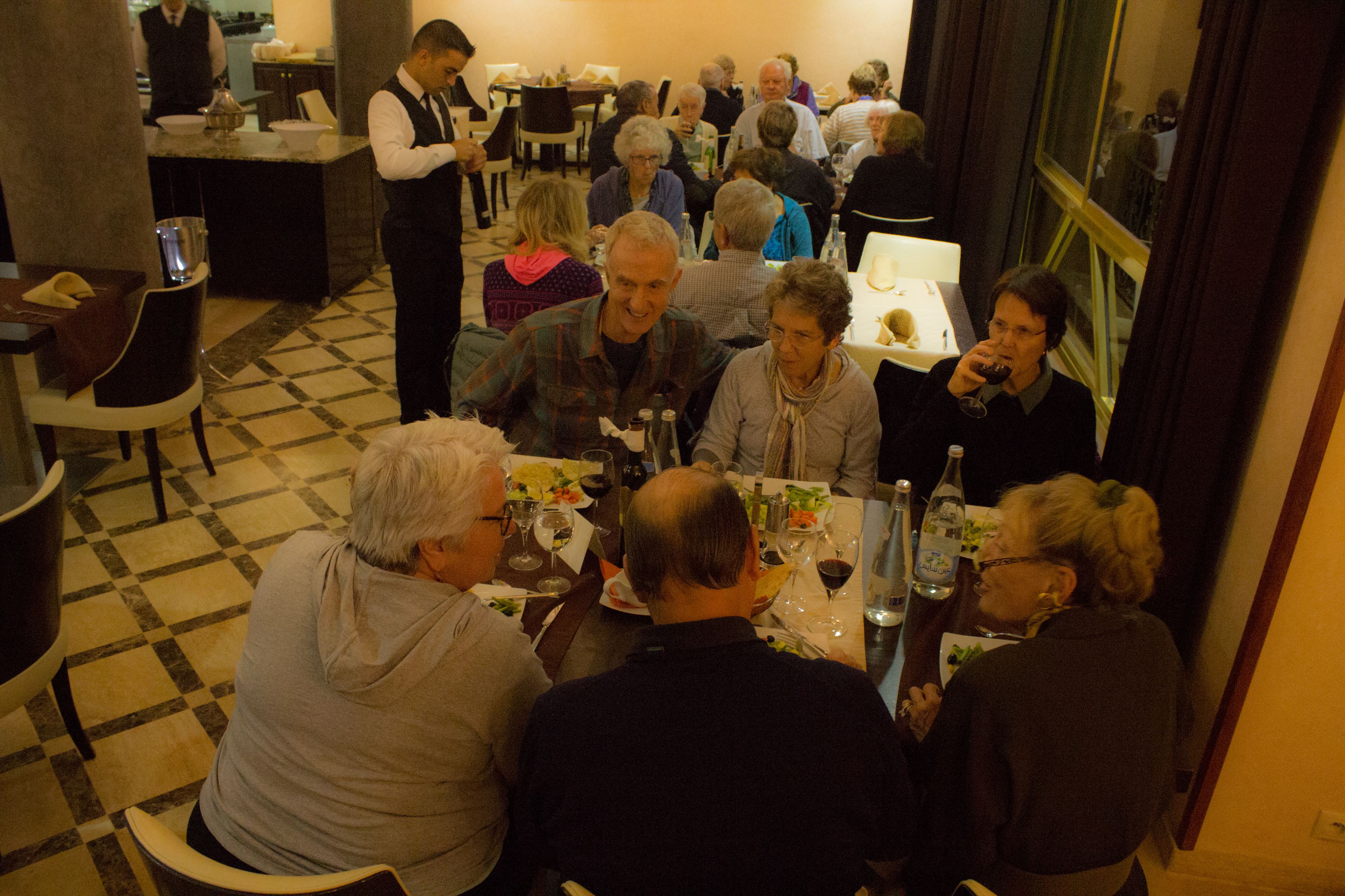 En gruppe mennesker spiser på en restaurant med et skakternet gulv, mens en tjener tager sig af dem.