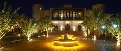 A courtyard with a fountain, palm trees, and a building with illuminated windows and steps.