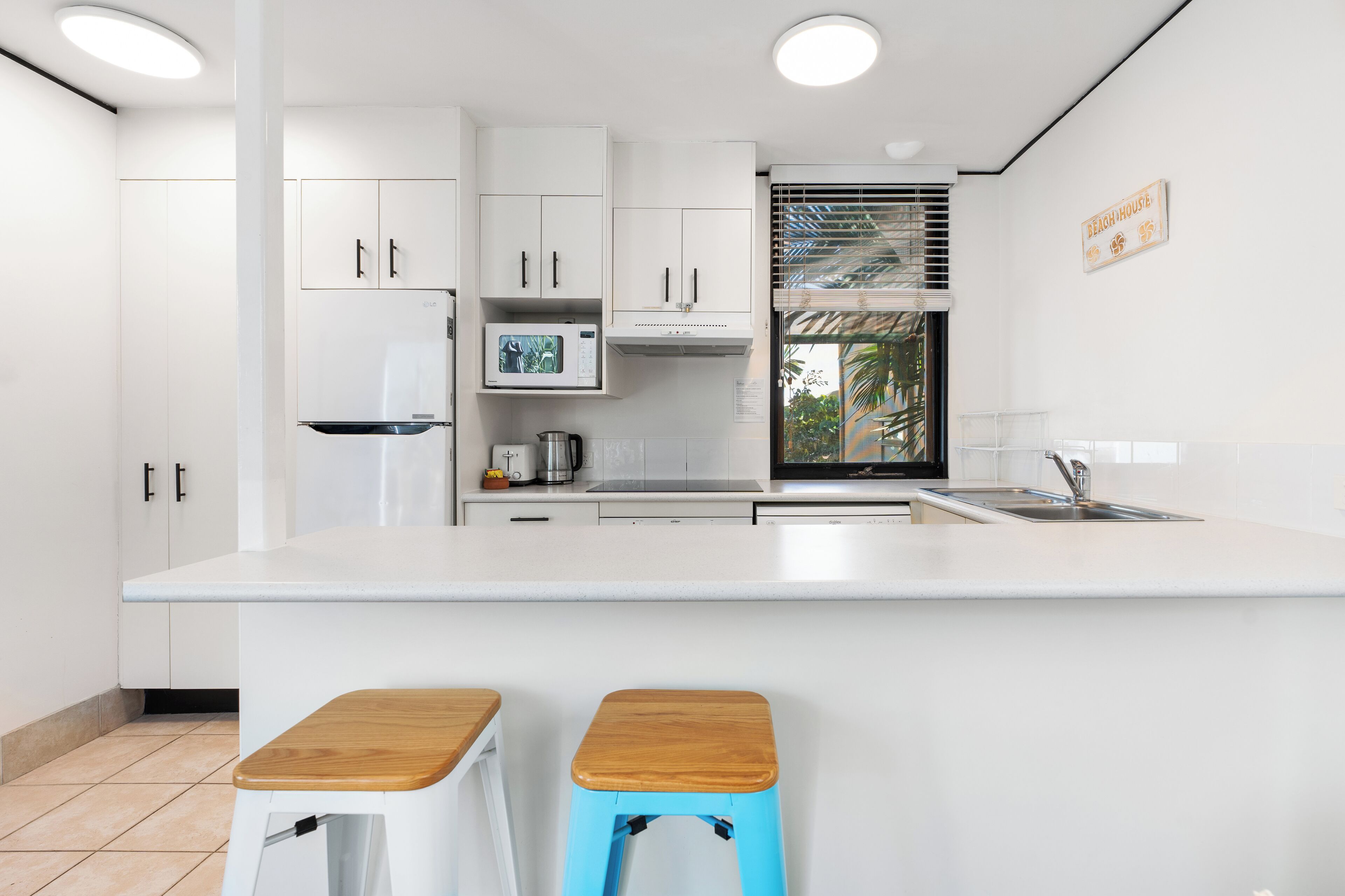 A modern kitchen with white cabinets, a large island, a microwave, and a sink. There are two stools with wooden tops.