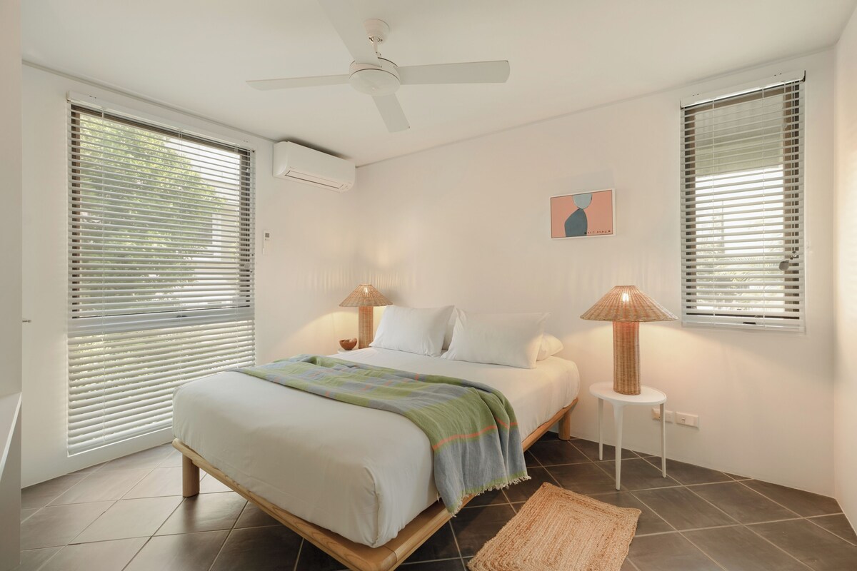 Une chambre à coucher avec un lit, deux lampes de chevet, un ventilateur de plafond et une fenêtre avec des stores.