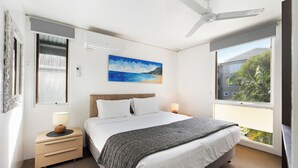 A bedroom with a bed, bedside tables, a ceiling fan, a painting of a beach, and a window with a view of greenery. - The Hastings Beach Houses (Noosa Heads)