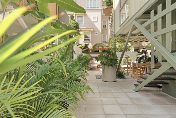 A narrow outdoor corridor with potted plants, a wooden table with chairs, and a staircase.