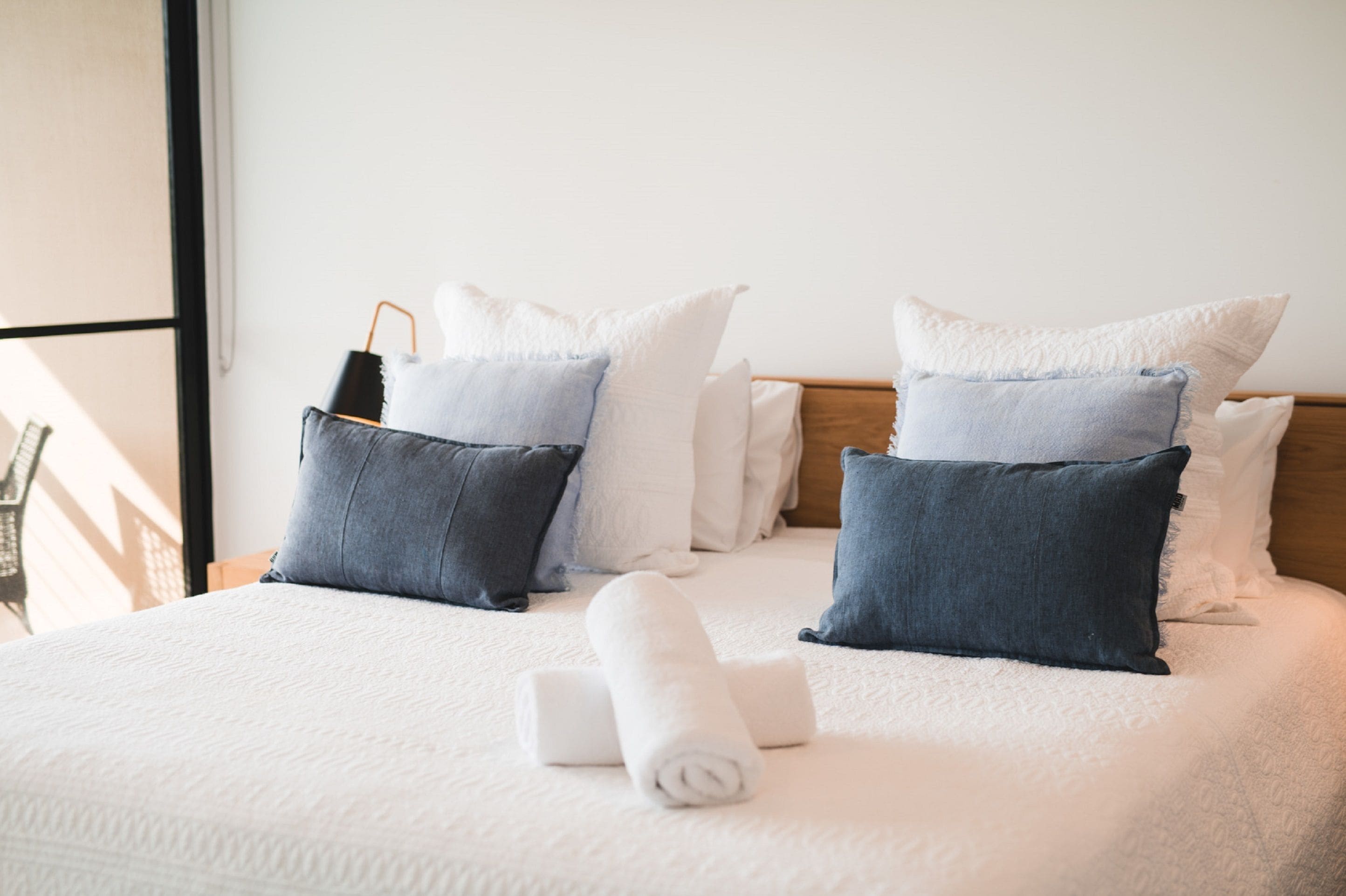 Een net opgemaakt bed met een witte deken, twee opgerolde handdoeken en een mix van witte en donkerblauwe kussens.
