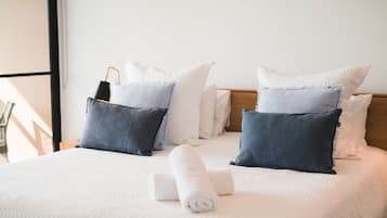 Een net opgemaakt bed met een witte deken, twee opgerolde handdoeken en een mix van witte en donkerblauwe kussens.