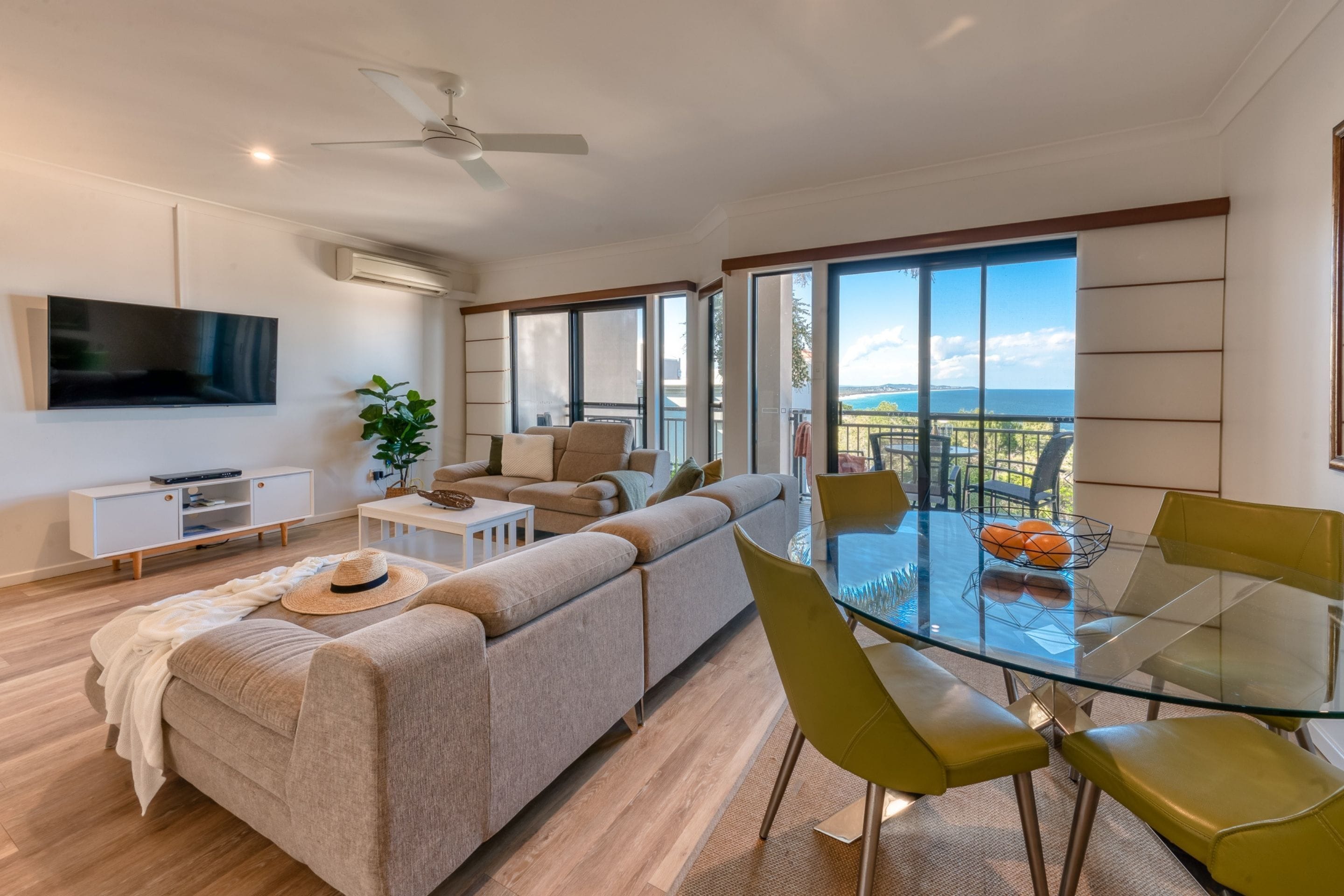 A modern living room with a sofa, a glass dining table with chairs, a TV mounted on the wall, and a large window offering a view of the sea.