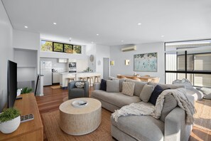 A modern living room with a sofa, coffee table, and a dining area with a kitchen in the background. - The Point Coolum Beach (Coolum Beach)