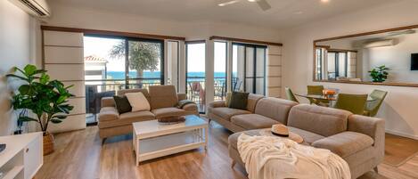 A modern living room with two sofas, a coffee table, and a dining area with a glass table and chairs. Large windows offer a view of the sea.