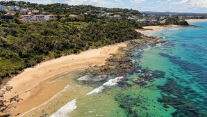 A coastal area with a sandy beach, clear turquoise waters, and a backdrop of residential buildings on a hillside. - The Point Coolum Beach (Coolum Beach)