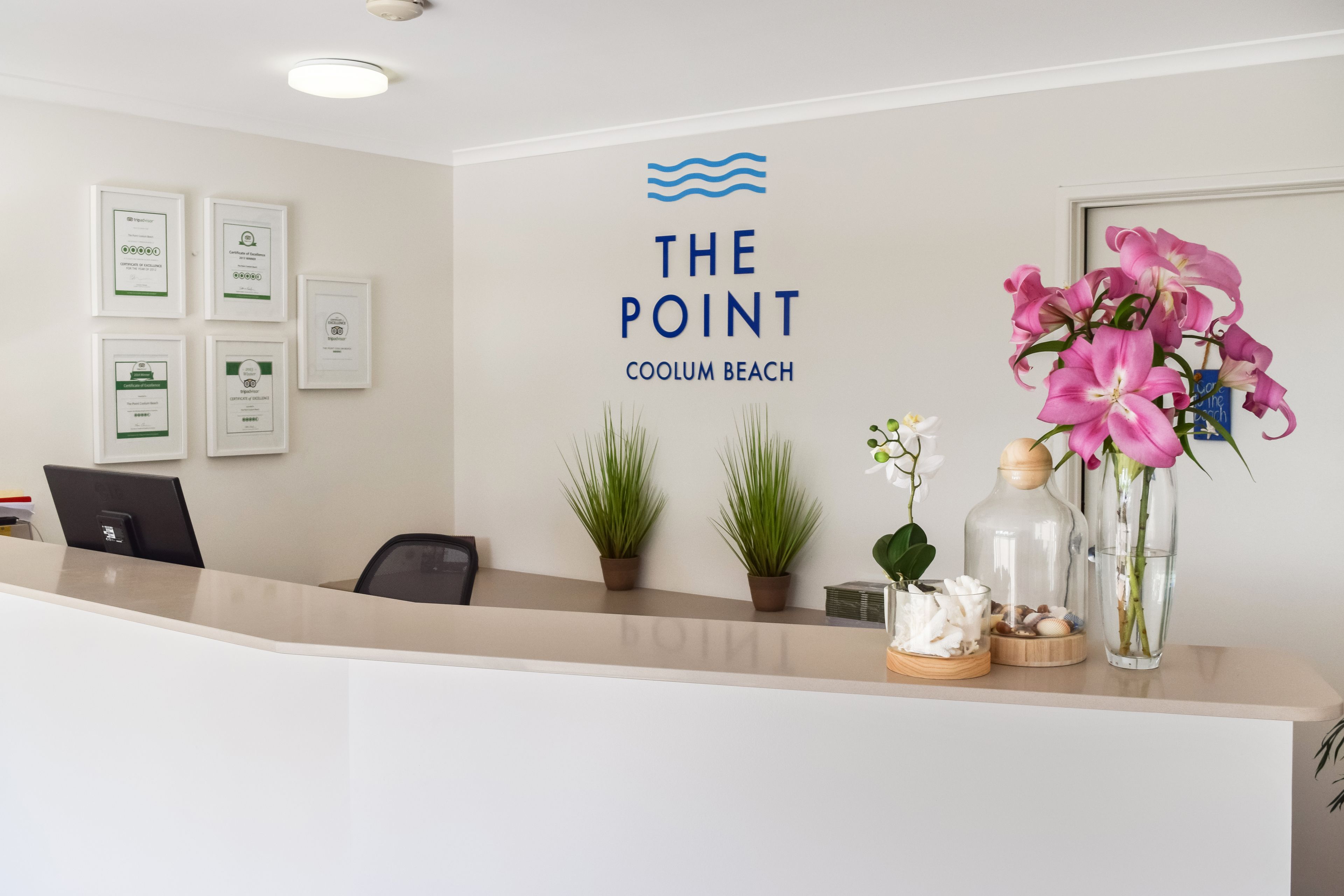 Un comptoir d’accueil orné du logo de « The Point Coolum Beach », des plantes en pot et un vase rempli de fleurs roses.